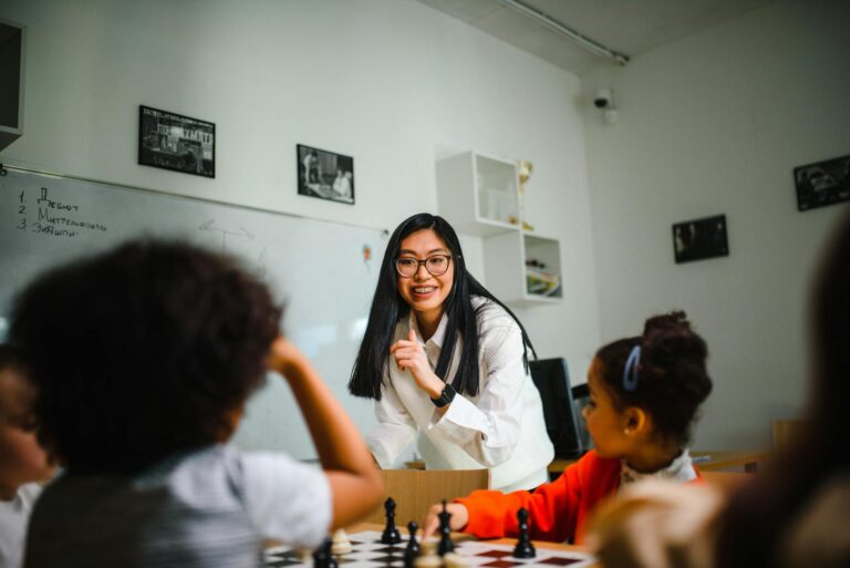 A teacher interacts with children during a chess lesson in a classroom setting.